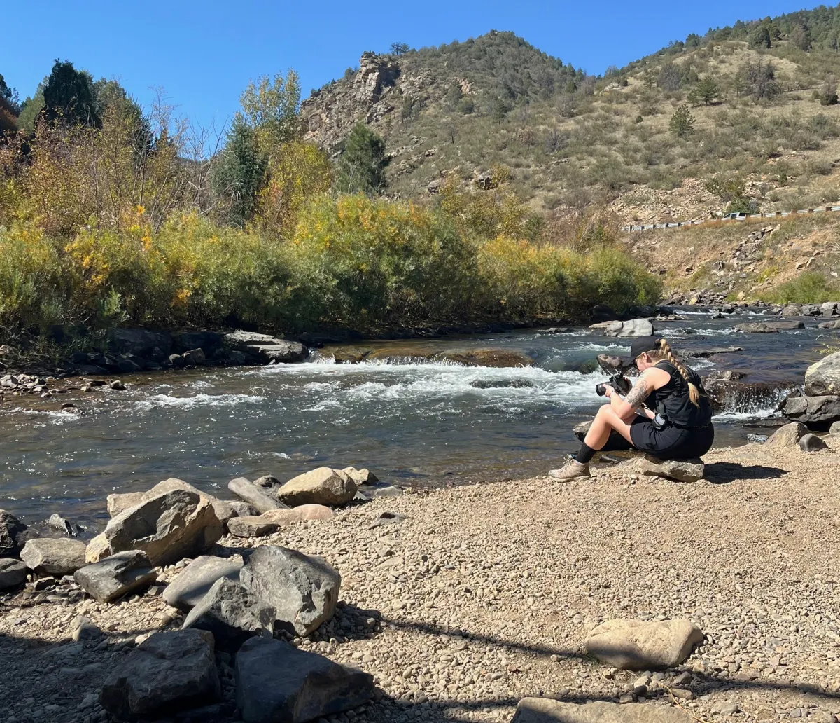 Mary-Ann Zykin sitting next to a flowing river with a camera taking photographs.